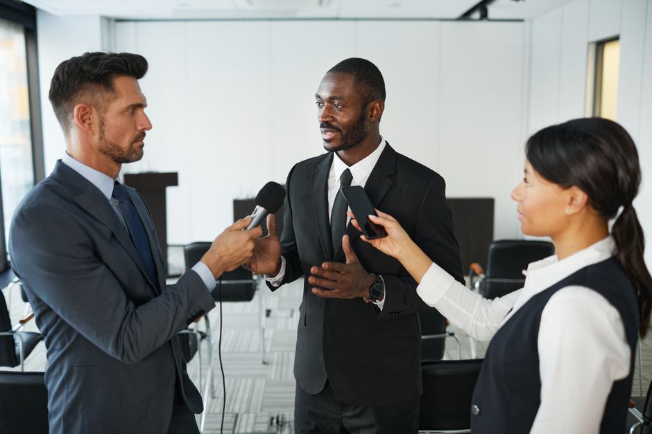 African American businessman in formal attire being interviewed by journalists with microphones in a conference room.